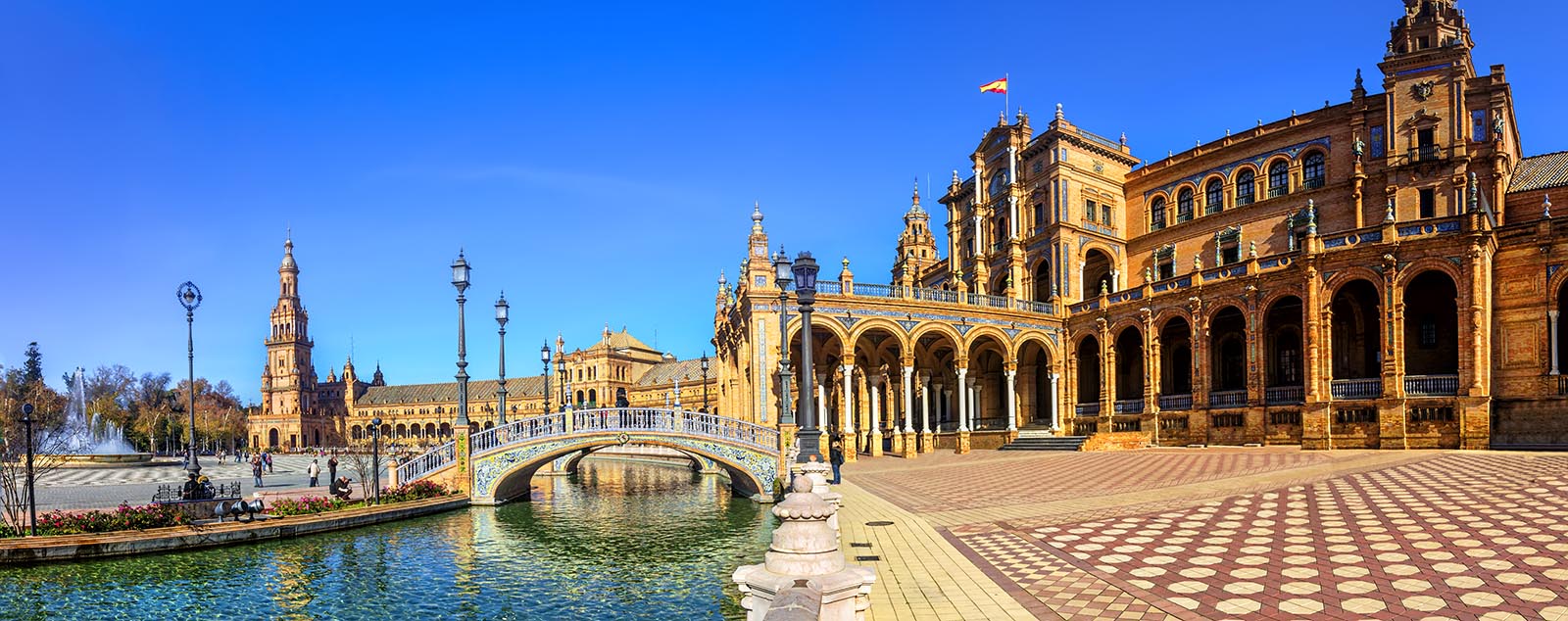 An ornate building with a row of arches, tiled bridges and blue canal on a bright sunny day, Plaza de España, Seville, Andalucia
