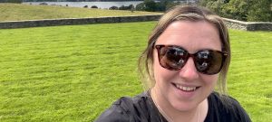 Amy Bassil takes a selfie picture in front of lush green fields and a lake surrounded by forests in the distance