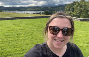 Amy Bassil takes a selfie picture in front of lush green fields and a lake surrounded by forests in the distance.