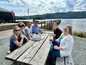 A small group are having refreshments outside Windermere Jetty Museum on a picnic bench next to Lake Windermere.