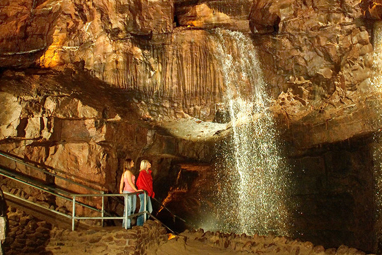 National Showcaves Centre of Wales, Cathedral cave Two people on a walkway inside a large cavern with a small waterfall cascading down the rockface in front of them