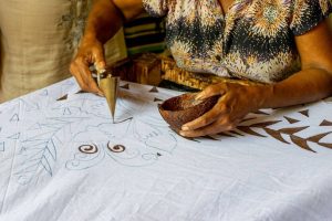 Close up of a worker outlining an intricate design on fabric with gold tool. She is taking the brown dye from a half coconut shell