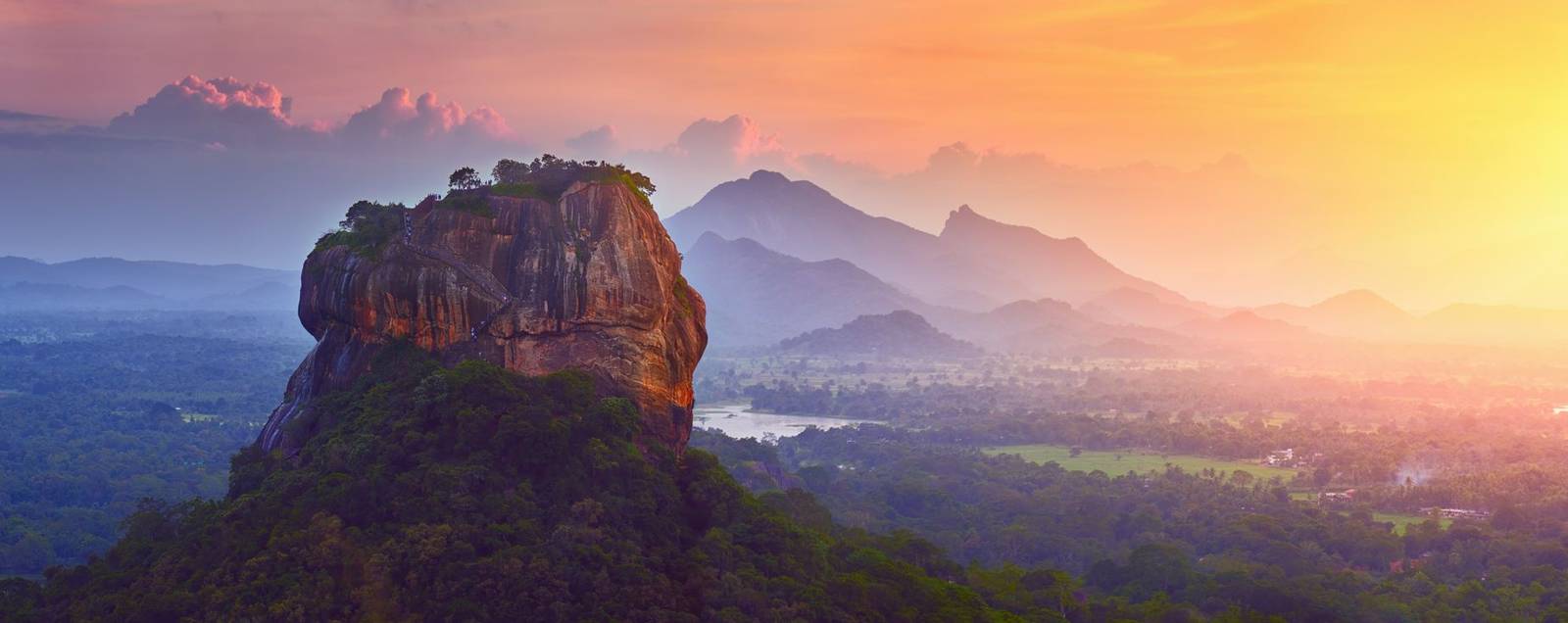 An aerial view of a large rock sitting at the top of a high ridge with a sea of forests below and shadows of mountain tops in the distance with the bright glow of a sunset to the right