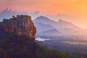 An aerial view of a large rock sitting at the top of a high ridge with a sea of forests below and shadows of mountain tops in the distance with the bright glow of a sunset to the right