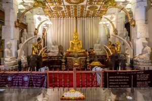 The inside of a buddhist temple with a central gold buddha and white and gold statues of buddha and elephant tusks all around. A flower offering is on the floor