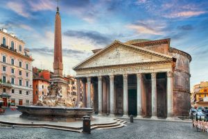 An ornate stone fountain in a cobbled square sits in front of a circular building with an ancient colonnaded facade (The Pantheon) set against a pink and blue clouded sky