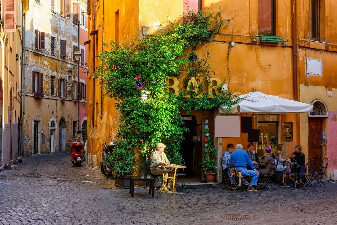 Italian cafe bar People sat on tables and chairs outside a bright yellow bar on a cobbled street with greenery growing over the walls