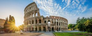 A circular four story ruin filled with arches on the first three floors (the Colosseum) and morning light shining through with speckled clouds in a blue sky