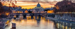 Lights are reflected in a river during dusk. A stone bridge of 5 arches spans the river and the domed basilica sits in the distance surrounded by other lit buildings