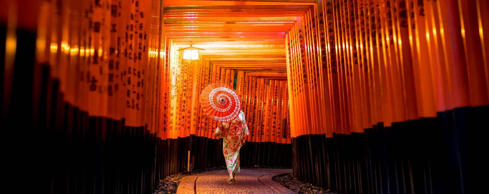 Fushimi Inari Shrine A woman dressed in a traditional silk kimono with a parasol walks under a row of tall red torii gates lined one after the other forming a red square tunnel