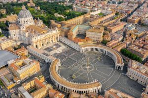 Aerial view of Vatican City and the expansive Saint Peter's Basilica with its grey-domed roof dominating the end of the huge circular colonnaded forecourt and square. Surrounding the grounds are various sized rectangular buildings of terracotta and cream.