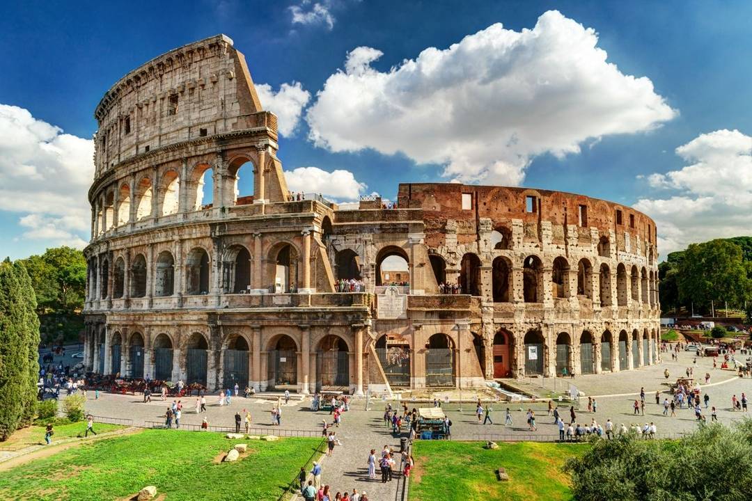 A circular four story ruin filled with arches on the first three floors (the Colosseum) with people on the path leading to it and on surrounding lawns.