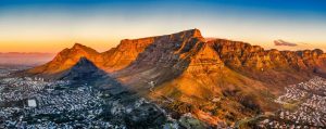 'Table Mountain', a large flat topped mountain glows orangey red in the sunset, with a large shadow cast over Cape Town below.