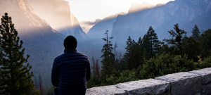 A man sits on a wall looking out into the distant landscape of pine trees and dramatic mountains during sunrise