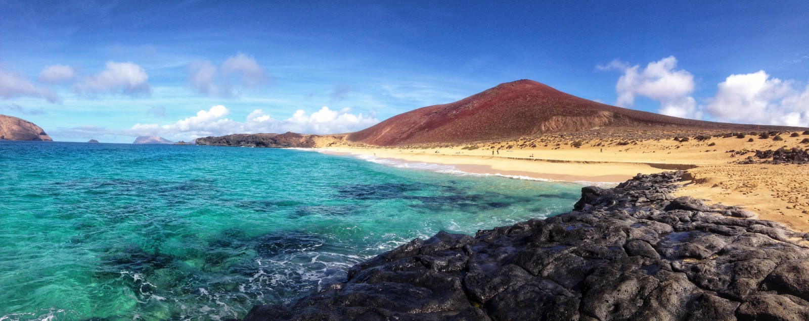 Turquoise waves roll against black volcanic rocks beside a long golden beach with a red volcano behind it and a bright blue sky