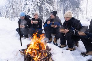 Travellers gather around an open fire to enjoy a hot meal in the snowy forest