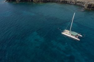 A catamaran with people on board sails along the cost with people swimming in the blue sea