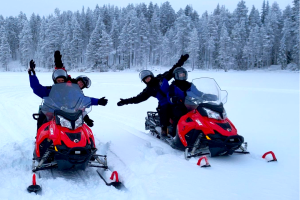 Two red snowmobiles with two travellers on each waving their hands in the air stand in a snowfield with snow-covered pines in the background