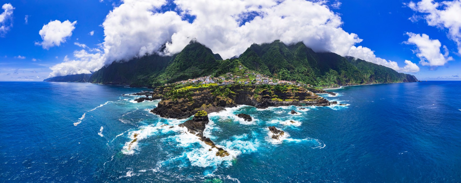 Panoramic view of a coastal village beneath cloud-covered green mountains, with Atlantic waves breaking against black volcanic rocks in Madeira