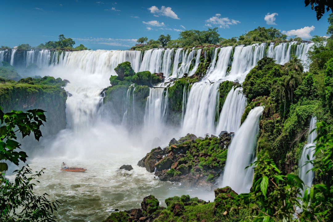 Wide view of Iguazú Falls cascading through lush tropical rainforest, with mist rising above the river and a small boat near the base of the waterfalls