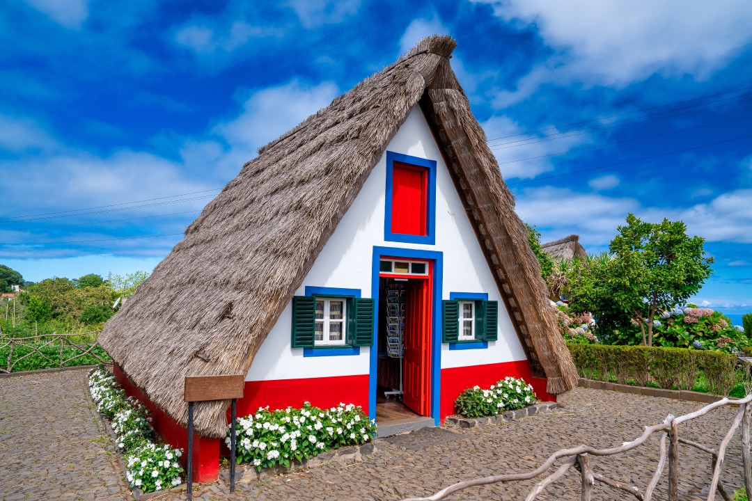 Traditional triangular thatched house with a steep straw roof, colourful painted doorway and flower beds in Santana