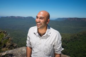 Image of Amar Latif looks off to the left smiling with blue skies and dramatic scenery of forests and mountains behind him. He's wearing a casual shirt and looks relaxed. 