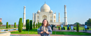 Leslie, a western woman with long brown tousled hair and wearing a floral dress, stands in front of the magnificent white marbled Taj Mahal with it's distinctive onion dome and manicured gardens. She holds her hands palms together in front of her heart and is smiling warmly.