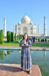 Leslie stands in a long Indian design dress, palms together in a prayer position with the white iconic Taj Mahal behind with its lush green manicured gardens and reflective waters.