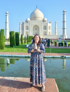 Leslie stands in a long Indian design dress, palms together in a prayer position with the white iconic Taj Mahal behind with its lush green manicured gardens and reflective waters.