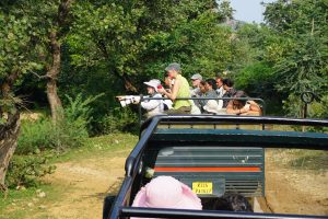 Leslie on safari in an open top cruiser with the group holds a long lens camera and points towards the forest