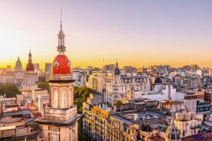 Panoramic view across the Buenos Aires skyline at sunset, with ornate historic towers, domes and city buildings glowing in warm evening light