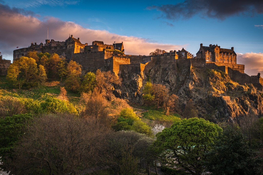 Edinburgh Castle rises above trees and rocky slopes in a warm evening light