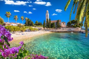 Franciscan Monastery beside a pebble beach and clear turquoise water framed by palm trees and flowers, Hvar, Croatia