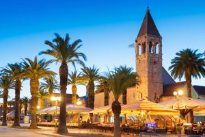 St Dominic Church and bell tower beside palm trees and outdoor cafés at dusk, Trogir, Croatia