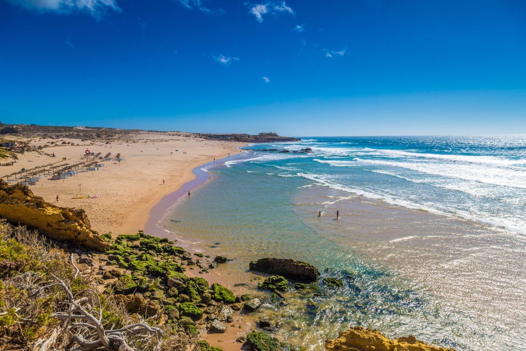 Praia Grande Beach Wide golden sandy beach beside the blue Atlantic Ocean, with waves breaking along the shoreline and rocky cliffs in the foreground at Praia Grande, Sintra
