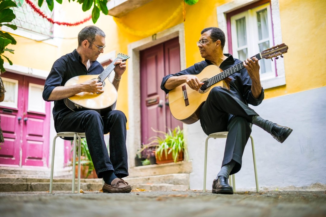 Two seated musicians playing a Portuguese guitar and an acoustic guitar in a courtyard with yellow walls and colourful doors in Lisbon